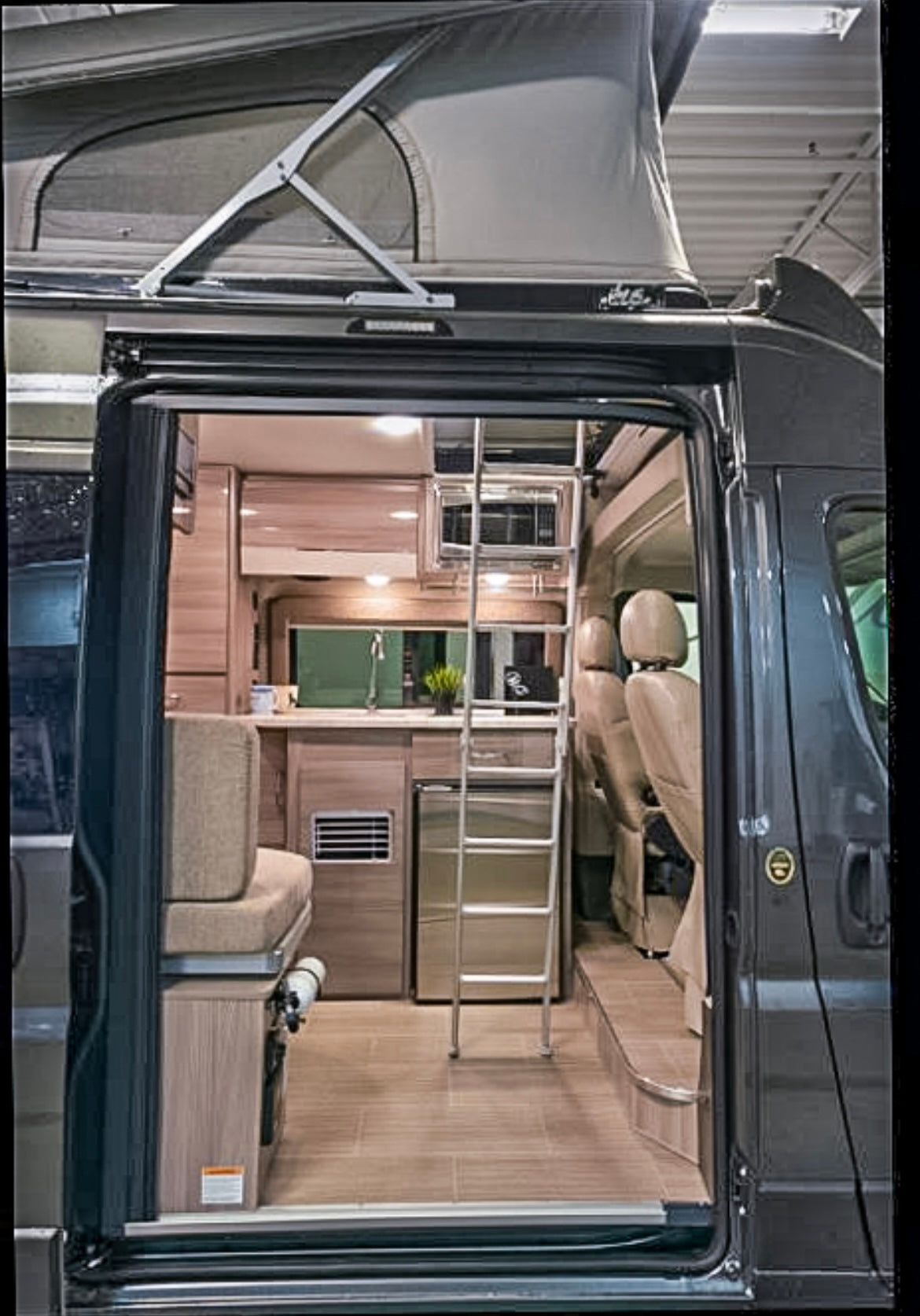 Explore this modern camper van RV rental interior! Features include a pop-top roof, kitchen, and ladder to an upper bed.