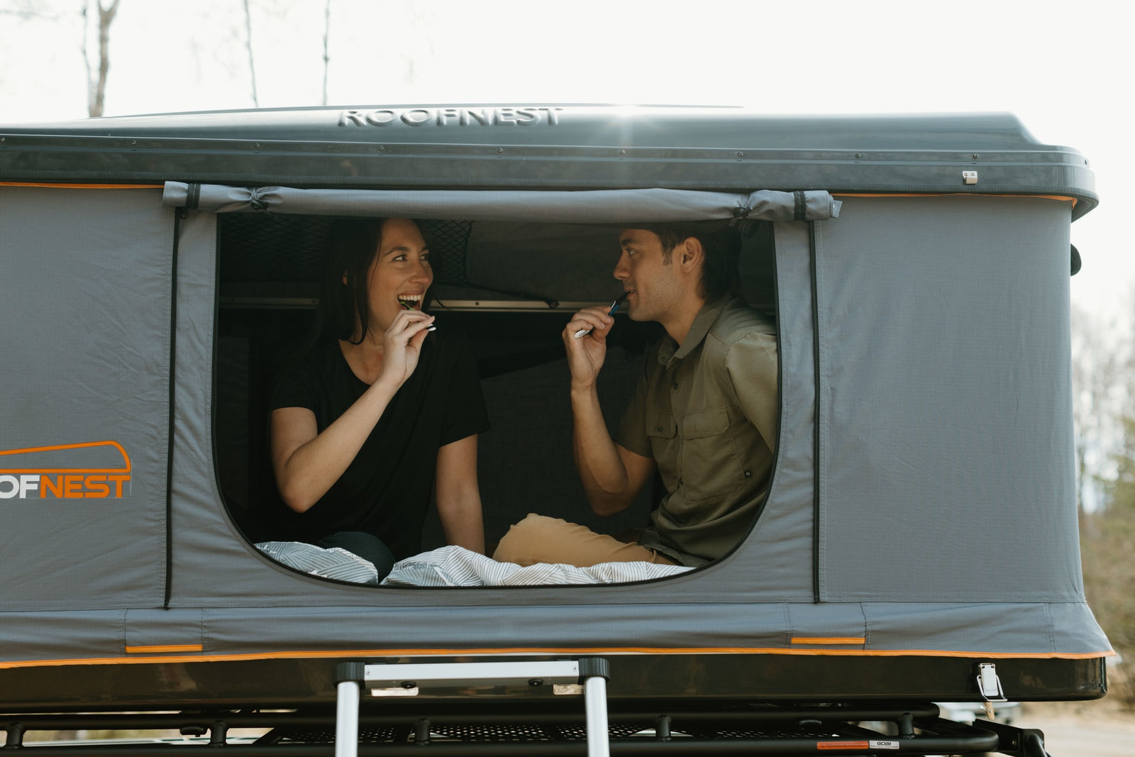 Adventure-ready RV rental alternative: Couple in a Roofnest roof-top tent brushing teeth, cozy camping outdoors.