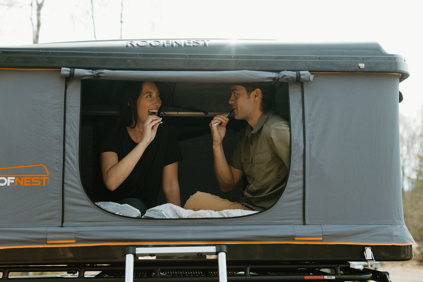 Adventure-ready RV rental alternative: Couple in a Roofnest roof-top tent brushing teeth, cozy camping outdoors.