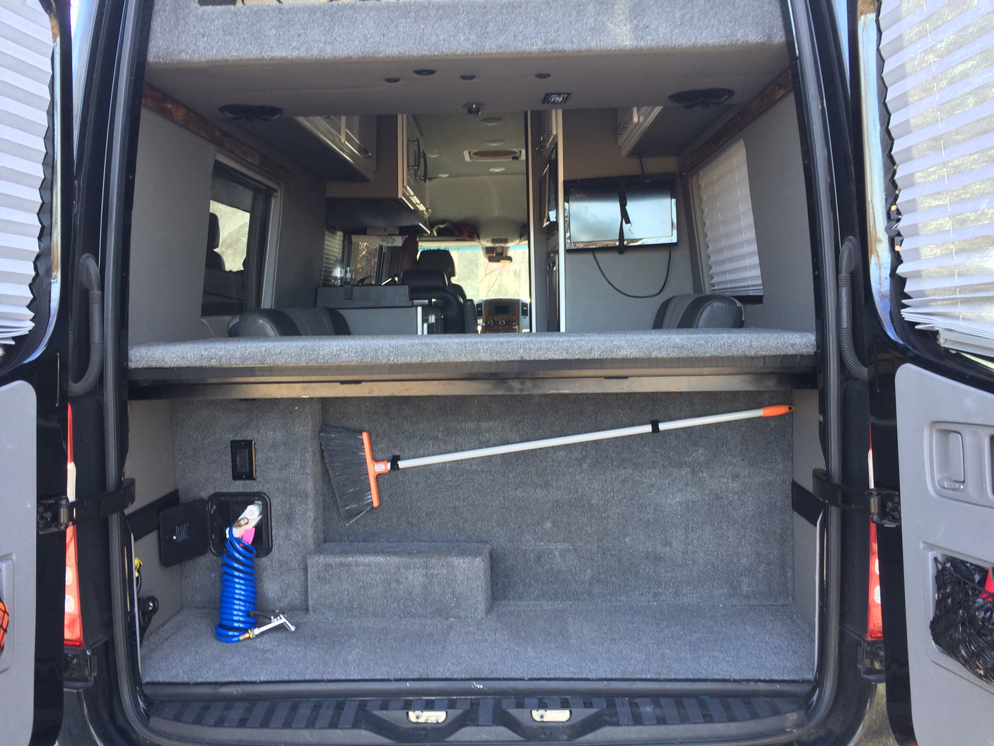 Spacious RV rental interior: two-tier setup with lower carpeted cargo area holding a broom and hose, and an upper bed/storage platform.