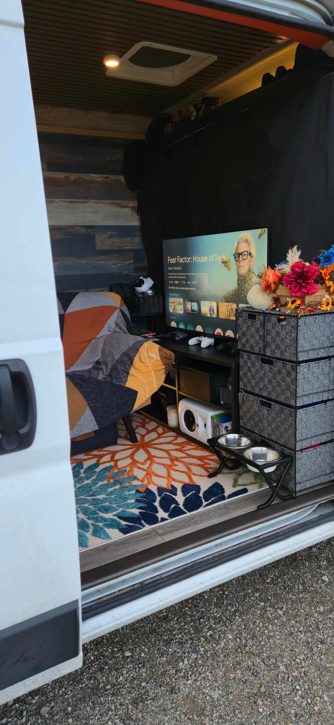 This rv rental offers a homey entertainment space with a TV showing "Fear Factor," gaming gear, pet bowls, & a comfy seat.