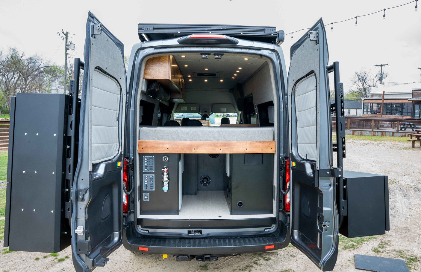 Custom RV rental van interior with open rear doors showing bed, utilities, and exterior storage boxes.
