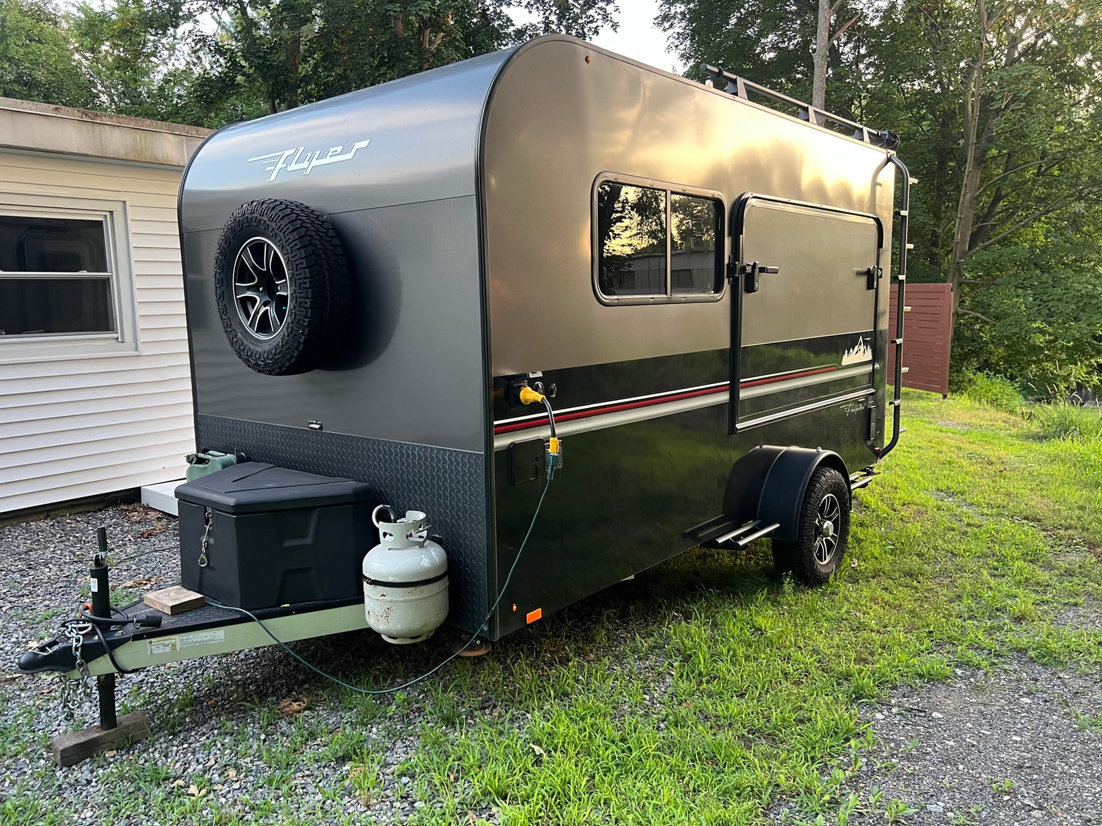 Dark gray "Flyer" teardrop RV rental, equipped with a spare tire & propane, parked on gravel for your next adventure.