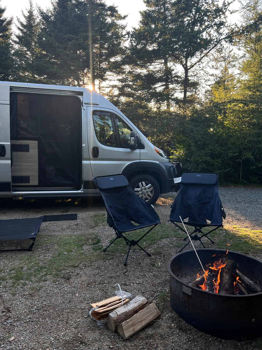 under blue sky & trees.|An RV rental at a forest campsite with two chairs & a burning campfire. Ideal for outdoor adventures & relaxation.|White camper van (RV rental) parked by a road in a vast