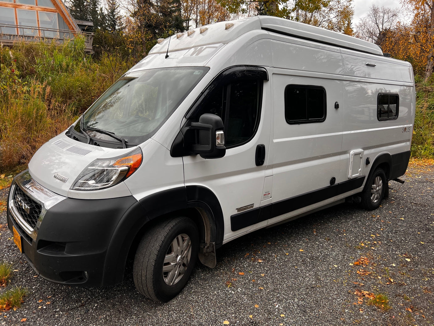 White Winnebago Solis camper van RV rental on gravel, surrounded by autumn foliage and a building.
