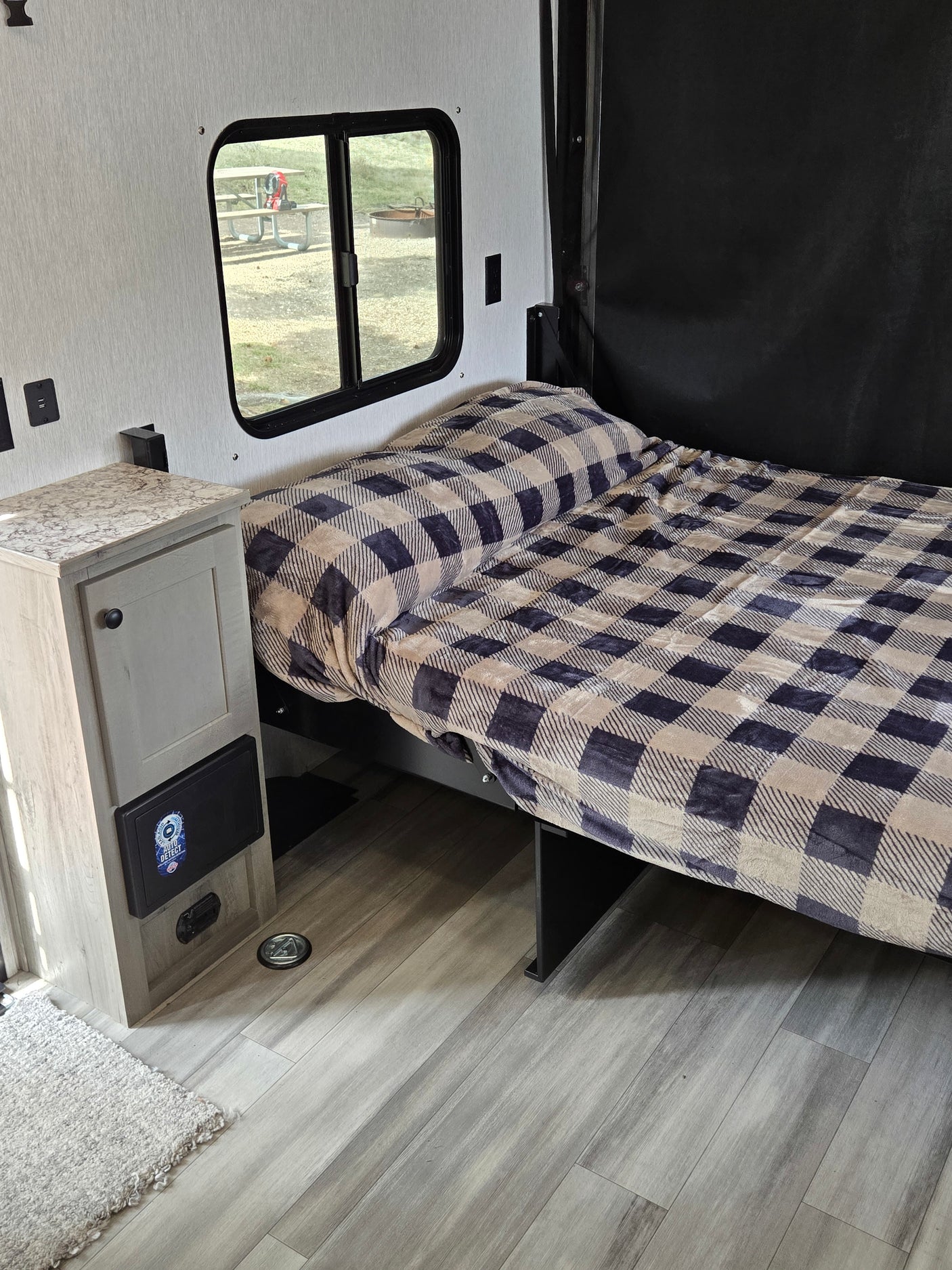 Cozy RV rental sleeping area: plaid bed, nightstand, and window view of a picnic area. USB outlets visible.