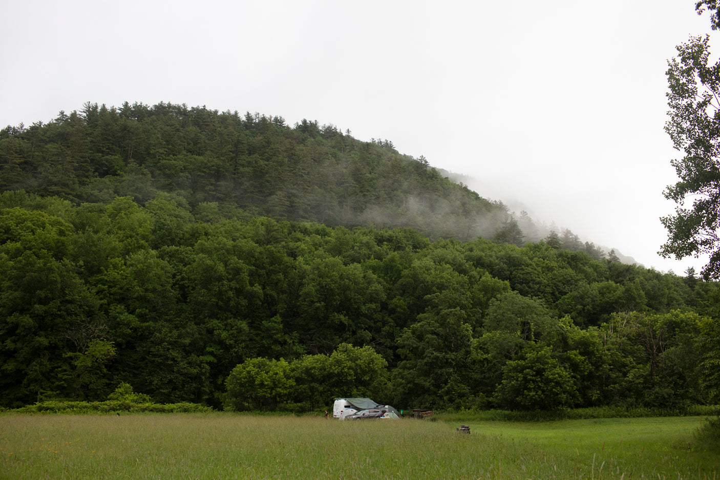 Experience nature with an RV rental! White RV & car parked in a lush green field before a misty, forested mountain.