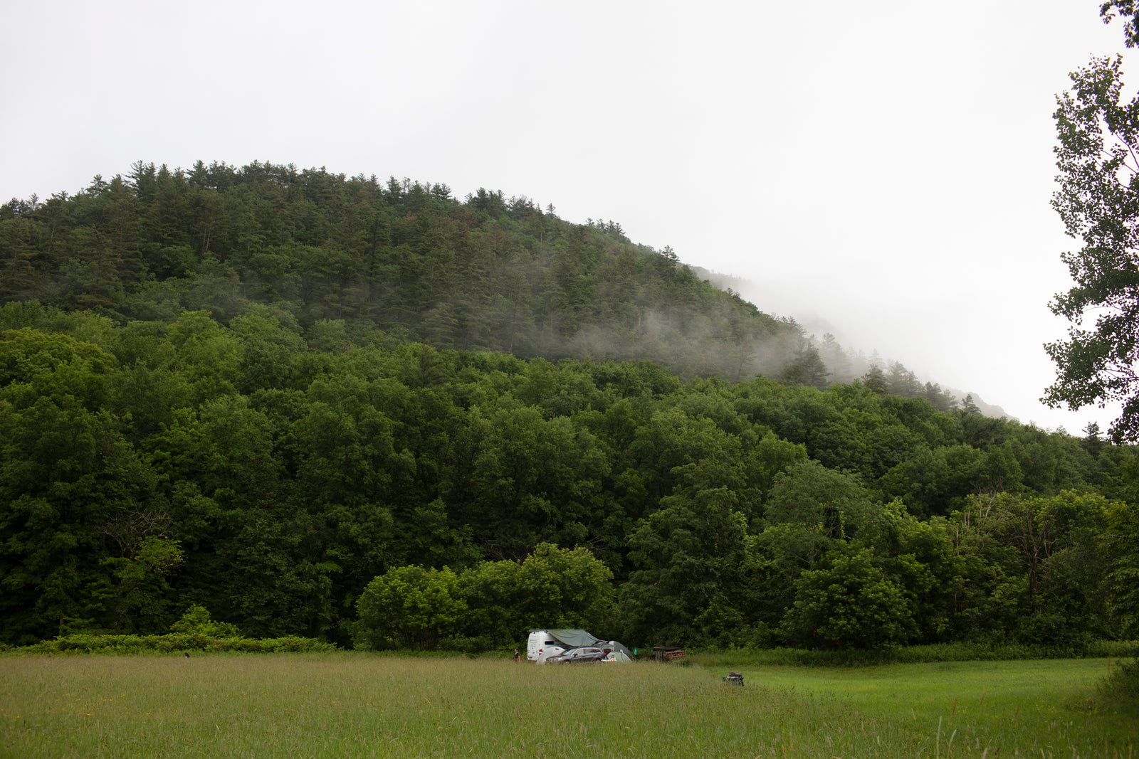 Experience nature with an RV rental! White RV & car parked in a lush green field before a misty, forested mountain.
