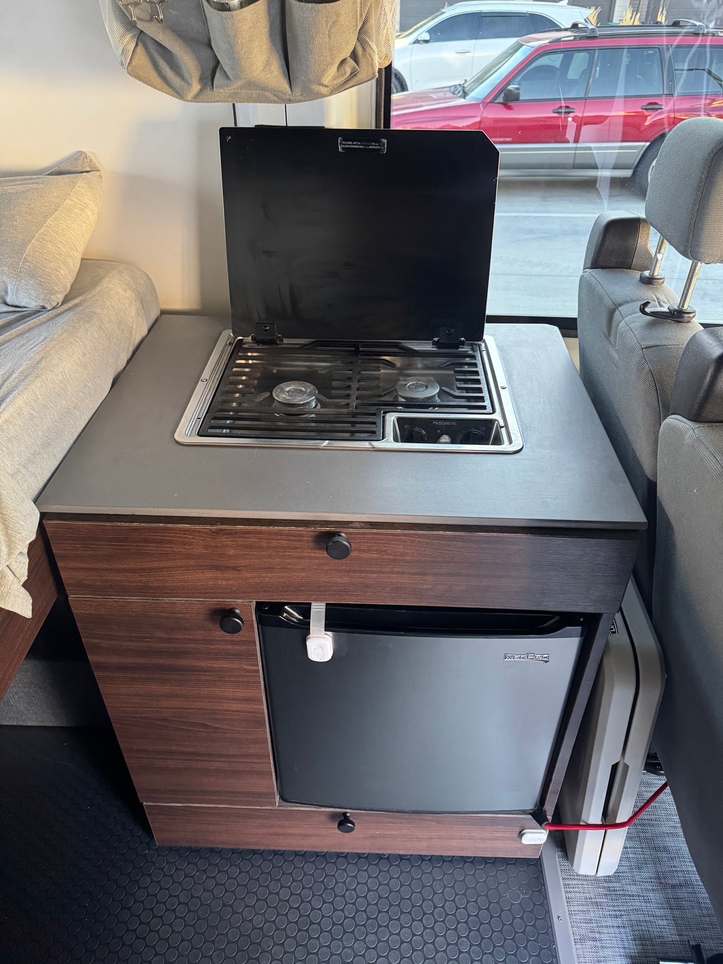 Compact RV rental kitchen with Dometic stove, sink, RedPro fridge, and adjacent bed, perfect for on-the-go meals and comfort.