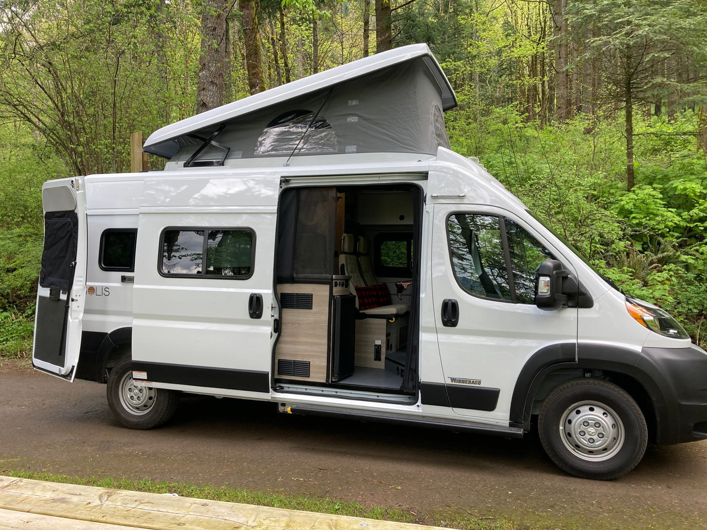 ready for your next RV rental adventure in nature.|White Winnebago Solis camper van RV rental parked in a lush forest campsite with chairs and fire pit.|Explore the outdoors in this white pop-top rv rental van with its side door open — photo 10