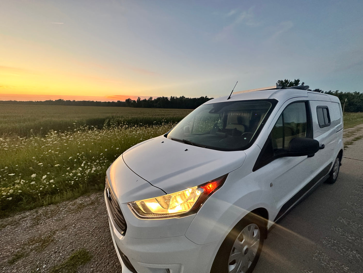 White camper van by wildflower field at sunset, headlights on. Great for an RV rental escape.