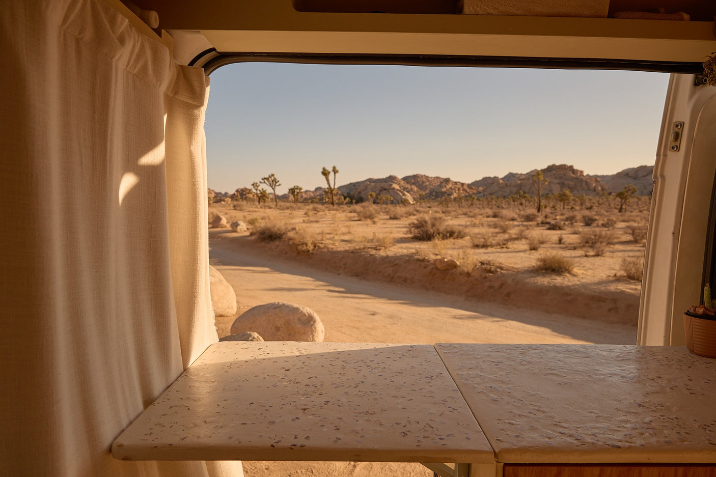 Inside an RV rental, a folding counter and curtains frame a window overlooking a desert road, Joshua trees, and distant mountains.