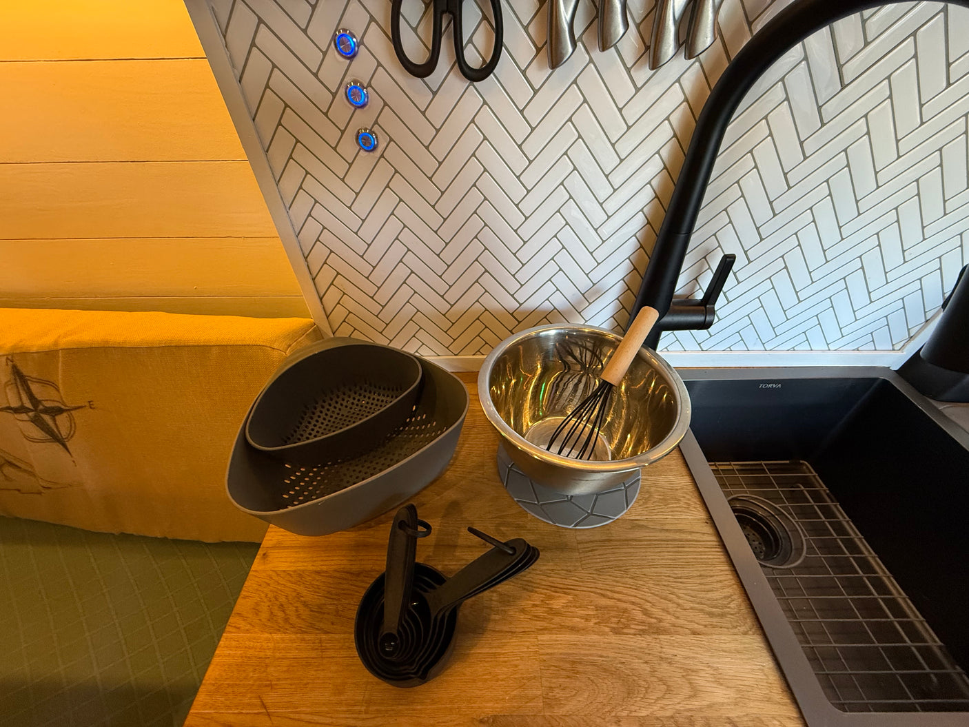 RV rental kitchen: wooden counter with black sink & faucet, herringbone backsplash, and cooking essentials like a colander, bowl, and measuring cups.