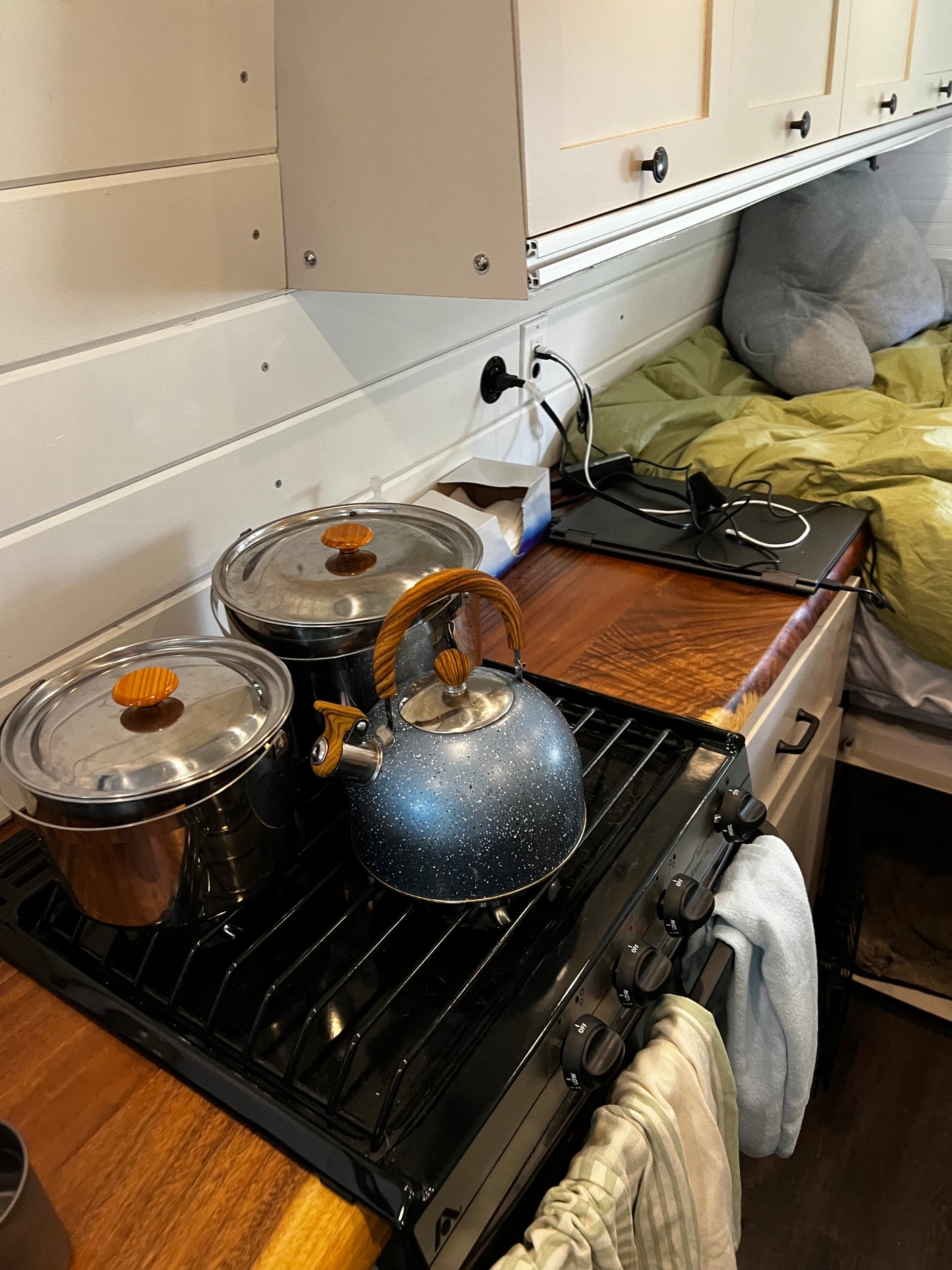 RV rental interior: A compact kitchen with stove, pots, and kettle, beside a sleeping area with a bed and laptop. — photo 7