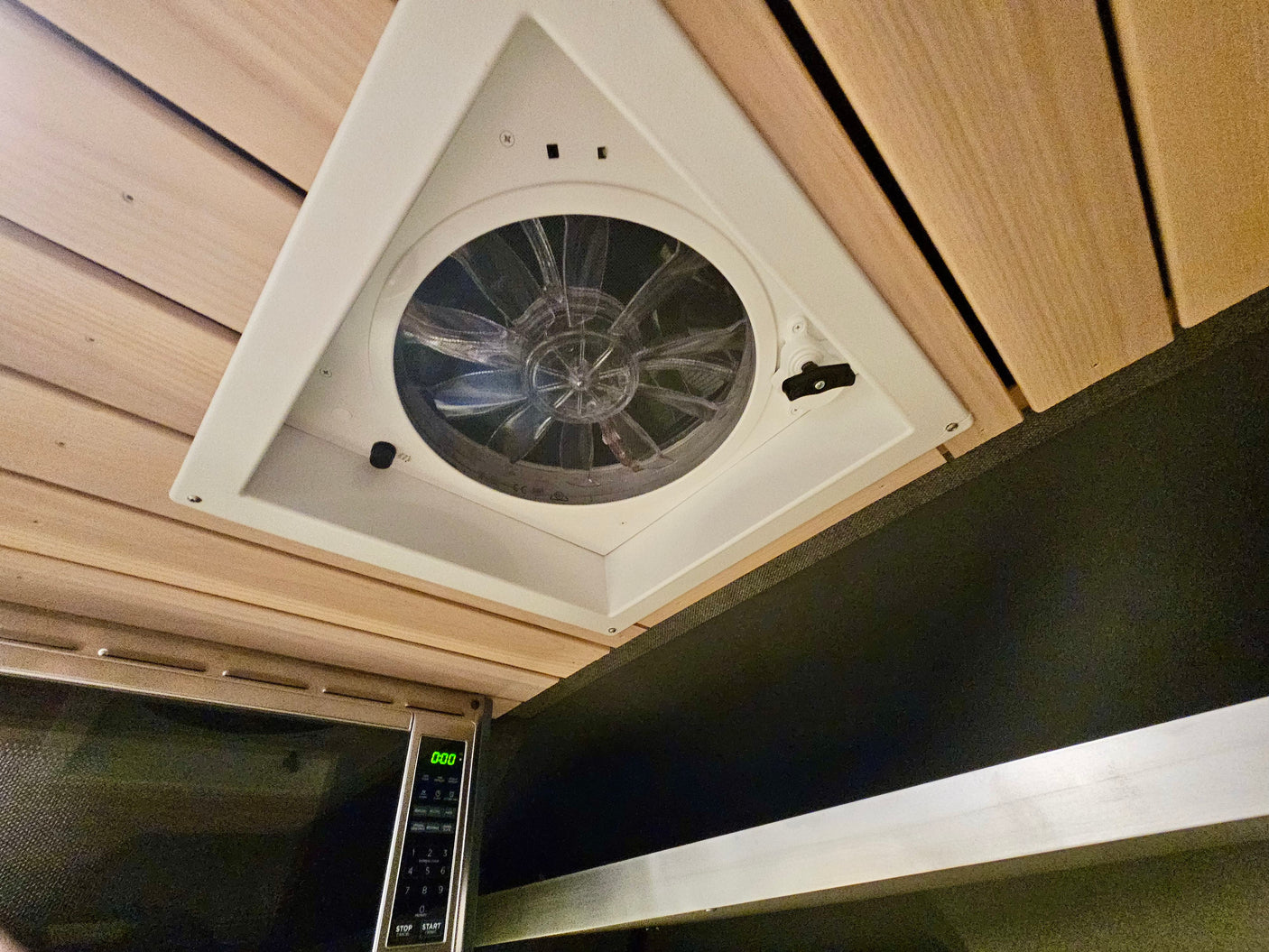 RV rental interior: A modern white ceiling vent fan with clear blades is centered, above a microwave and warm wood paneling.