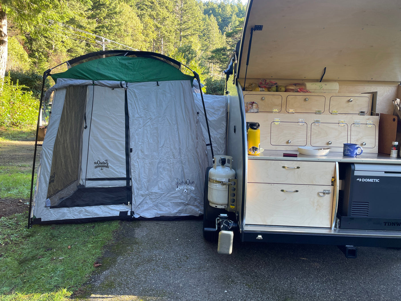 An RV rental setup shows an open camper kitchen next to a spacious tent, amidst green trees for outdoor camping.