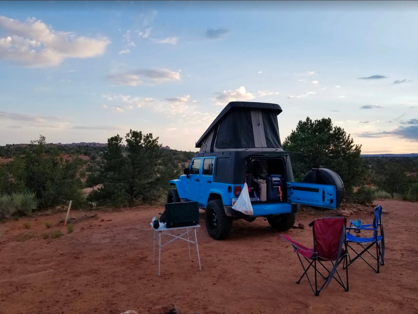 Overland camping at dusk! This blue pop-top Jeep with a stove & chairs makes for an ideal RV rental adventure.|Adventurous RV rental: A bright blue Jeep with a pop-top tent and a man
