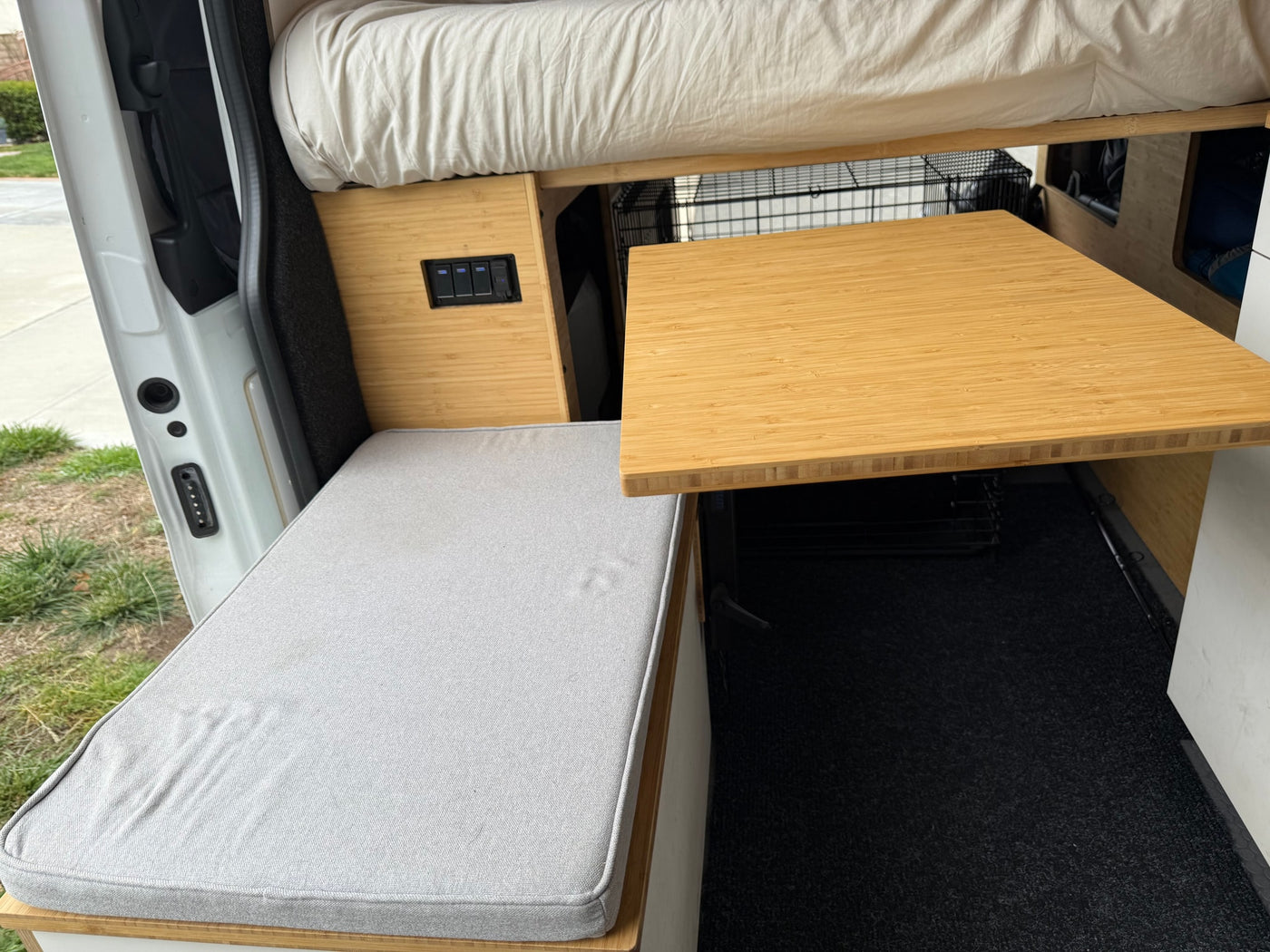 RV rental interior: bamboo table, cushioned bench, overhead bed, and pet crate. Controls visible by the open side door. — photo 16