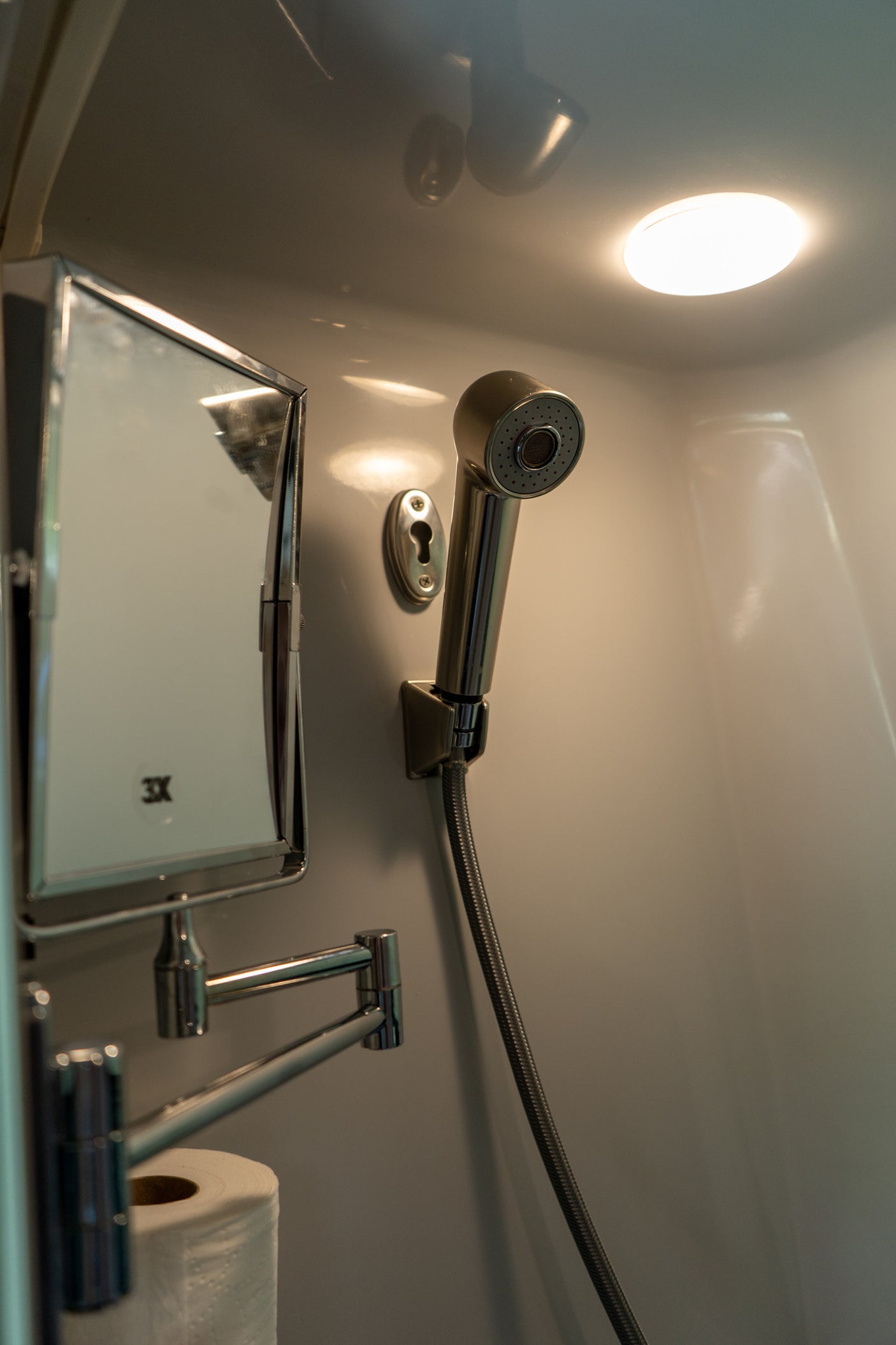 This RV rental's compact bathroom shows a hand-held shower, a multi-angle mirror, and toilet paper, all brightly lit from above.