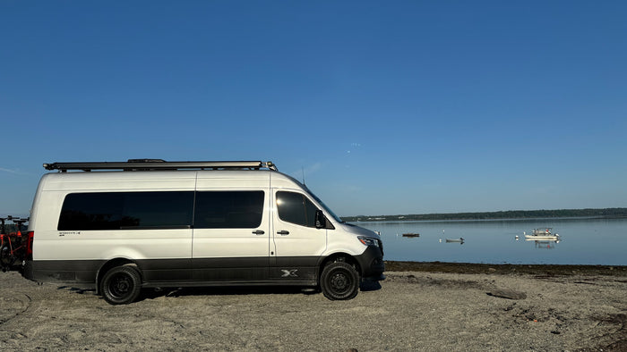 bikes ready for lakeside adventures.|White RV rental van with bikes on a gravel beach. Peaceful bay scene with boats & distant treeline under blue sky.|Silver Airstream RV rental camper van with bikes on a rack