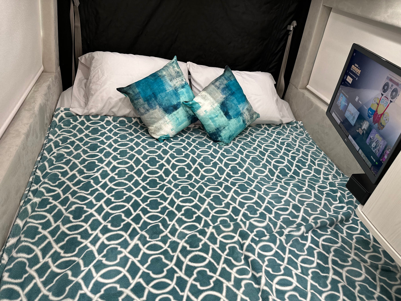 This RV rental features a cozy bed with a teal blanket, patterned pillows, & a TV playing Minions for a relaxing stay.
