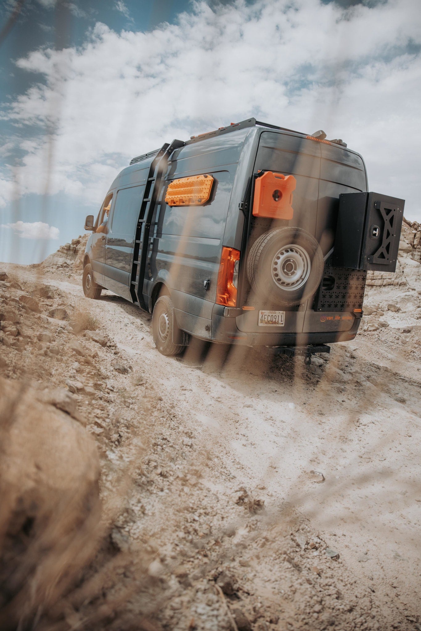 and fishing rod. Enjoy beach camping and outdoor adventures.|Off-road adventure in an RV rental: a customized dark gray Sprinter van ascends a dusty trail in a desert landscape.|Adventure-ready grey RV rental van with rear-mounted spare tire