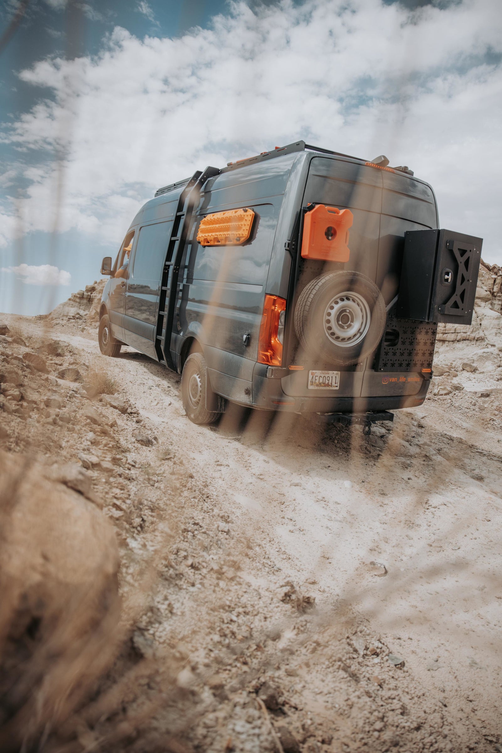 and fishing rod. Enjoy beach camping and outdoor adventures.|Off-road adventure in an RV rental: a customized dark gray Sprinter van ascends a dusty trail in a desert landscape.|Adventure-ready grey RV rental van with rear-mounted spare tire