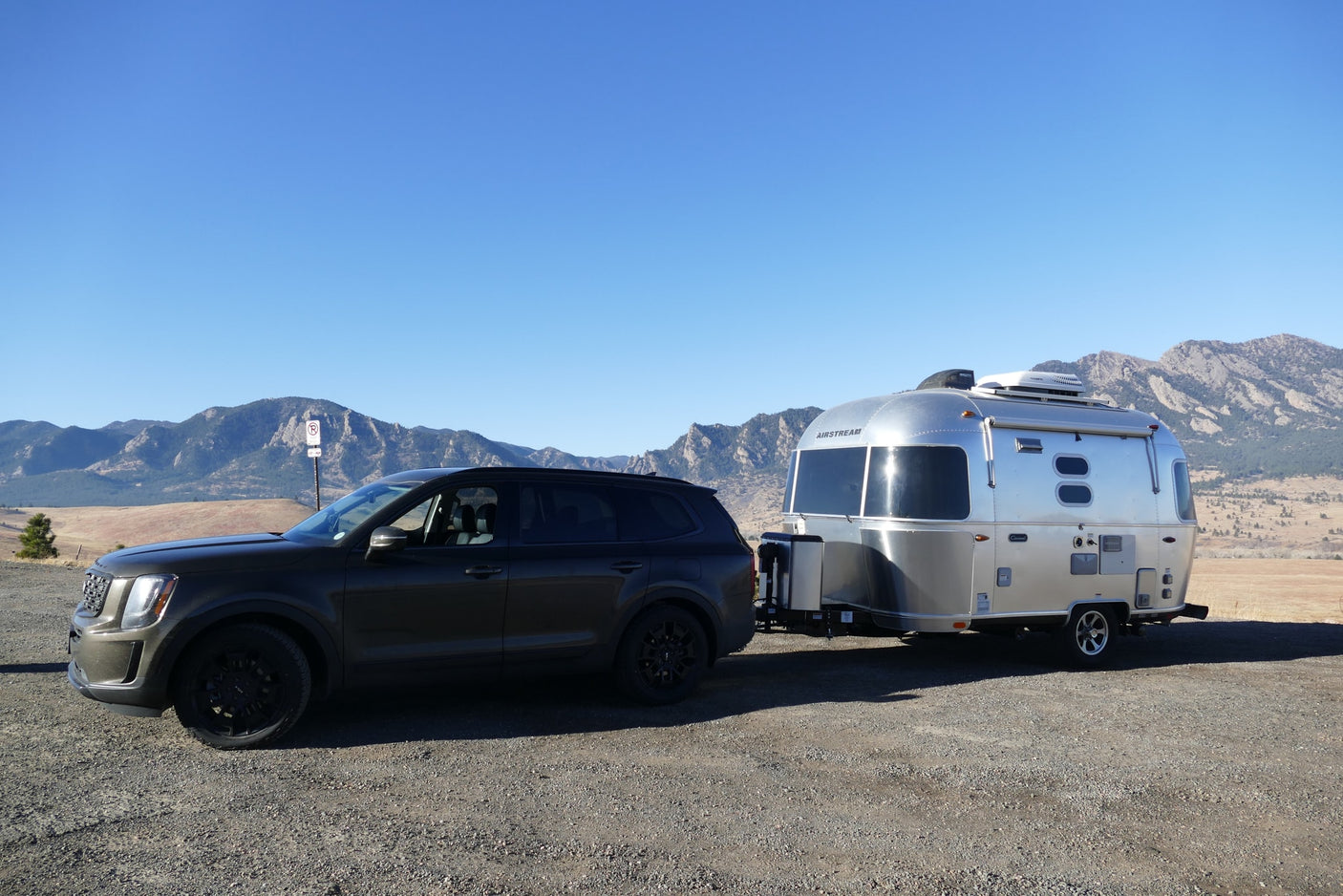 Dark SUV towing a shiny Airstream rv rental on gravel. Mountains and clear blue sky in background, perfect for a travel adventure.