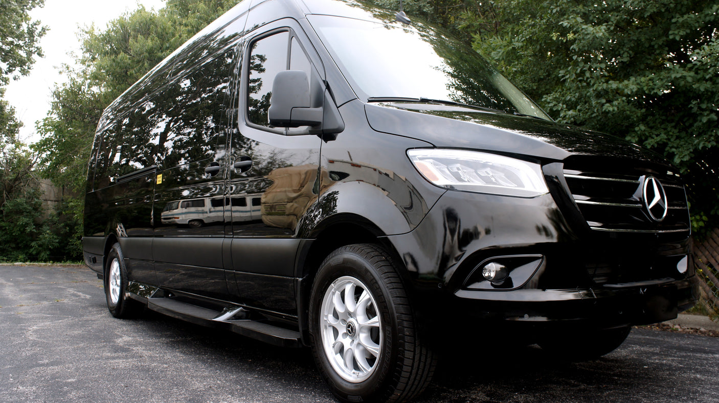 A sleek black Mercedes-Benz Sprinter van, ideal for a luxury trip or RV rental, reflecting trees and another vehicle.