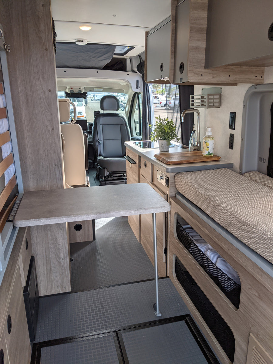 Inside a modern rv rental, featuring a compact kitchen, foldable table, and cozy bed area, looking towards the front.