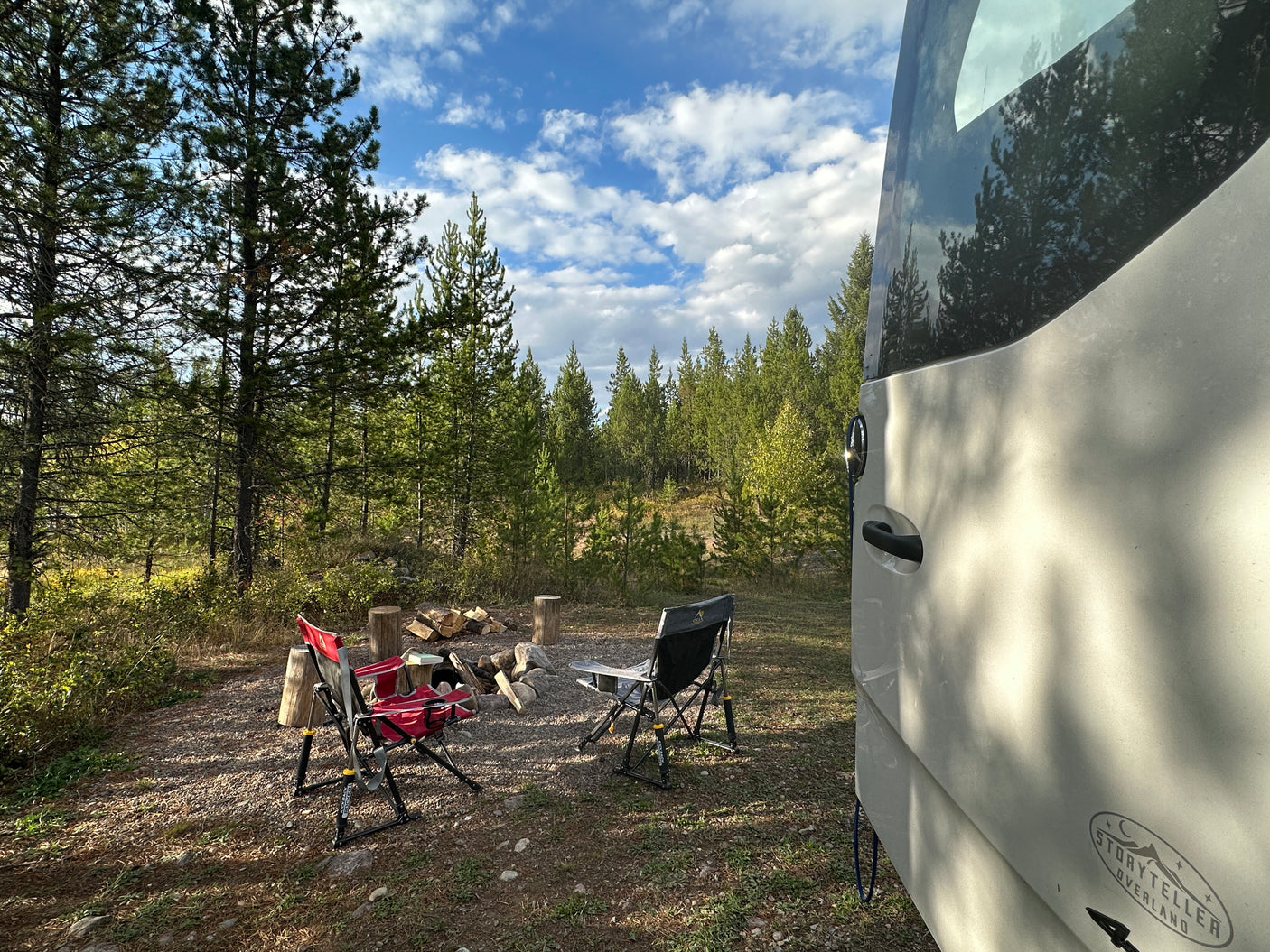 RV rental at a serene forest campsite, featuring camping chairs around a fire pit amidst tall pine trees under a blue sky.