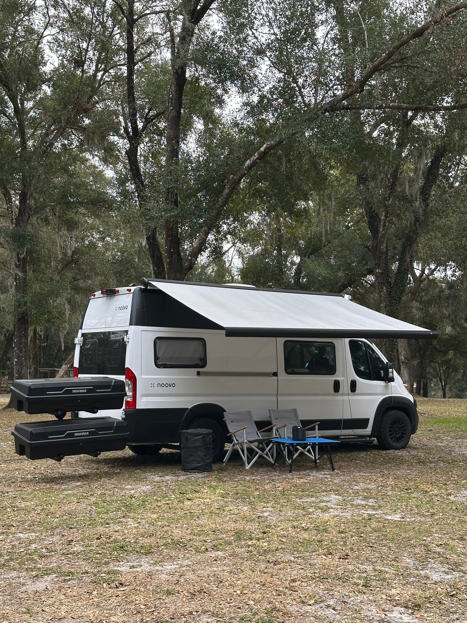White x noovo rv rental camper van with awning, chairs, and storage boxes. Set up for camping among mossy trees on a grassy outdoor site.