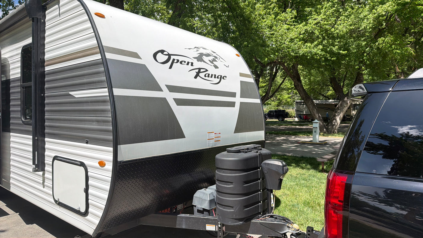 Front of an Open Range travel trailer, a great rv rental option, hitched to a black SUV in a sunny, green RV park.
