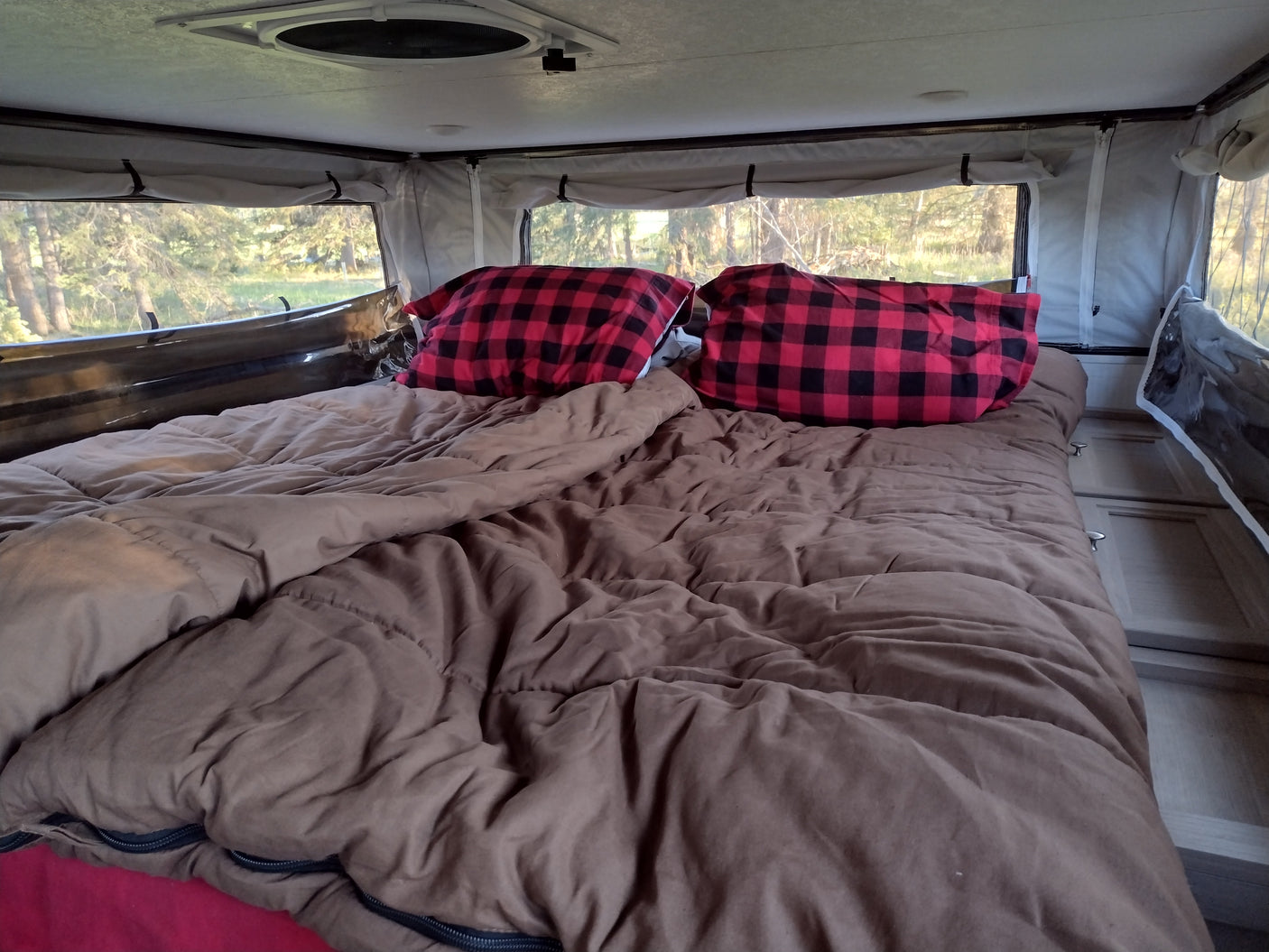 Inside a cozy RV rental: a comfortable bed with red plaid pillows and large windows showcasing forest views.