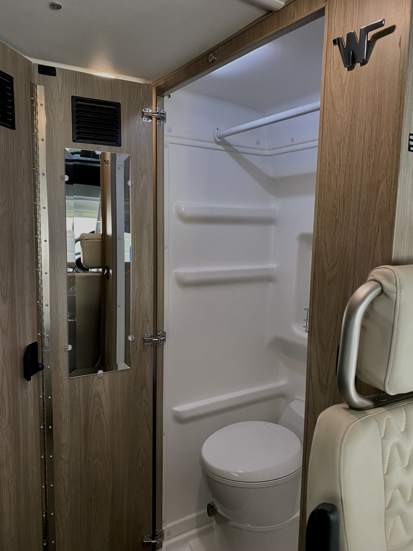 RV rental interior: a compact bathroom with a shower, toilet, and mirror, adjacent to a beige leather seat.