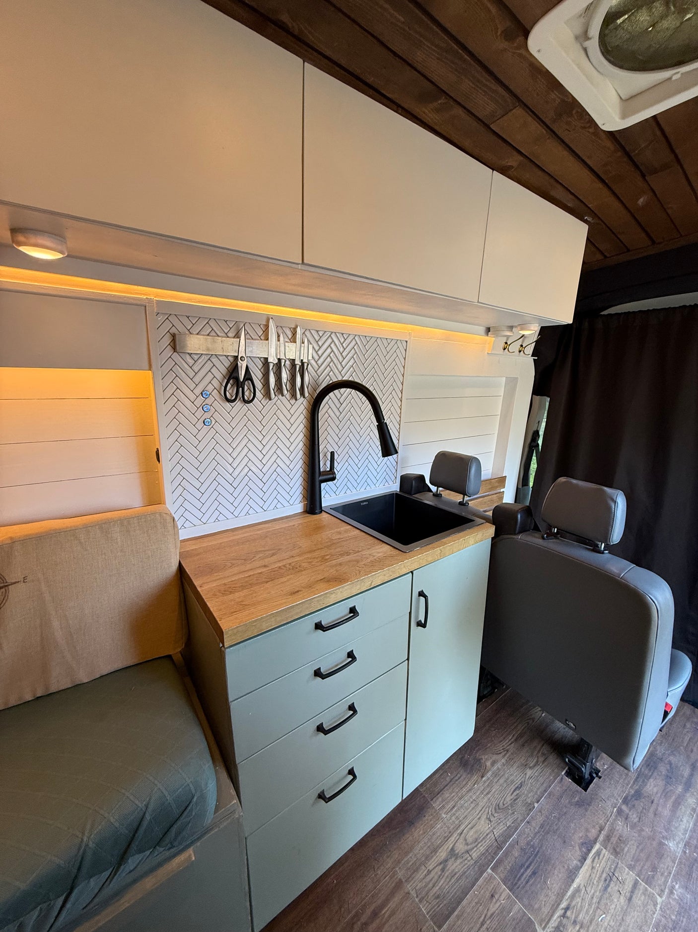 RV rental: Modern camper van kitchen with wood accents, a black sink, light gray cabinetry, and cozy seating.
