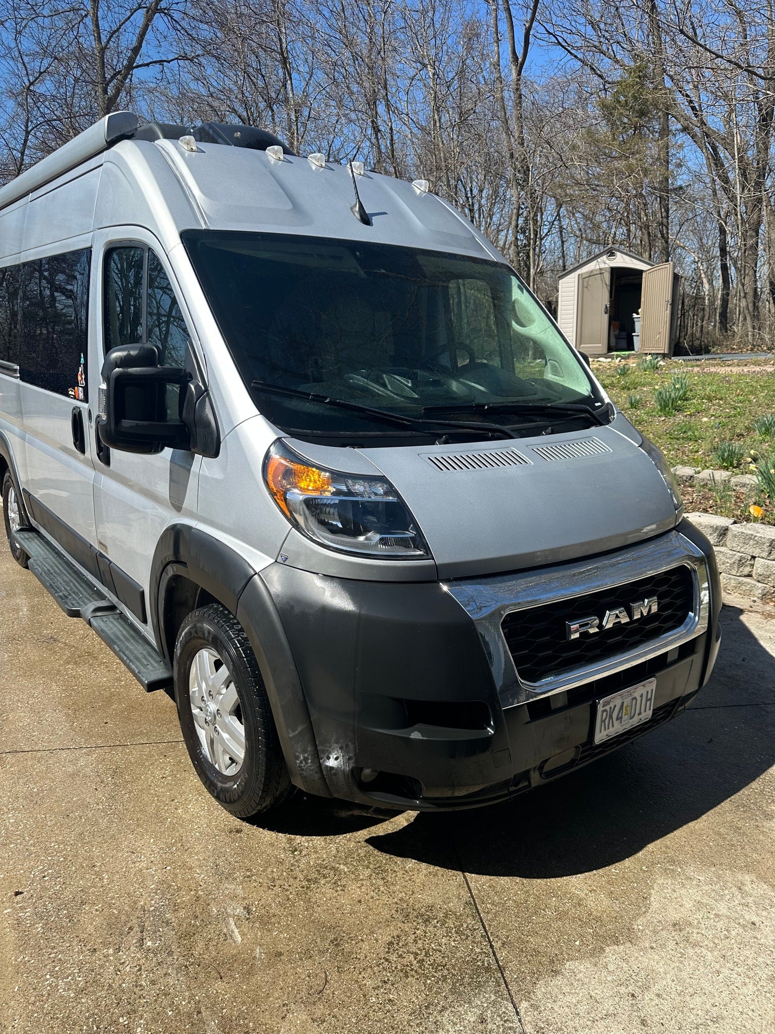 Silver RAM ProMaster RV rental, ready for adventure. Parked on a driveway, with a shed and trees in the background.