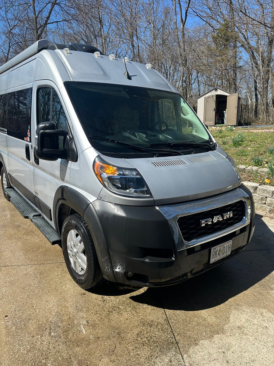 Silver RAM ProMaster RV rental, ready for adventure. Parked on a driveway, with a shed and trees in the background.