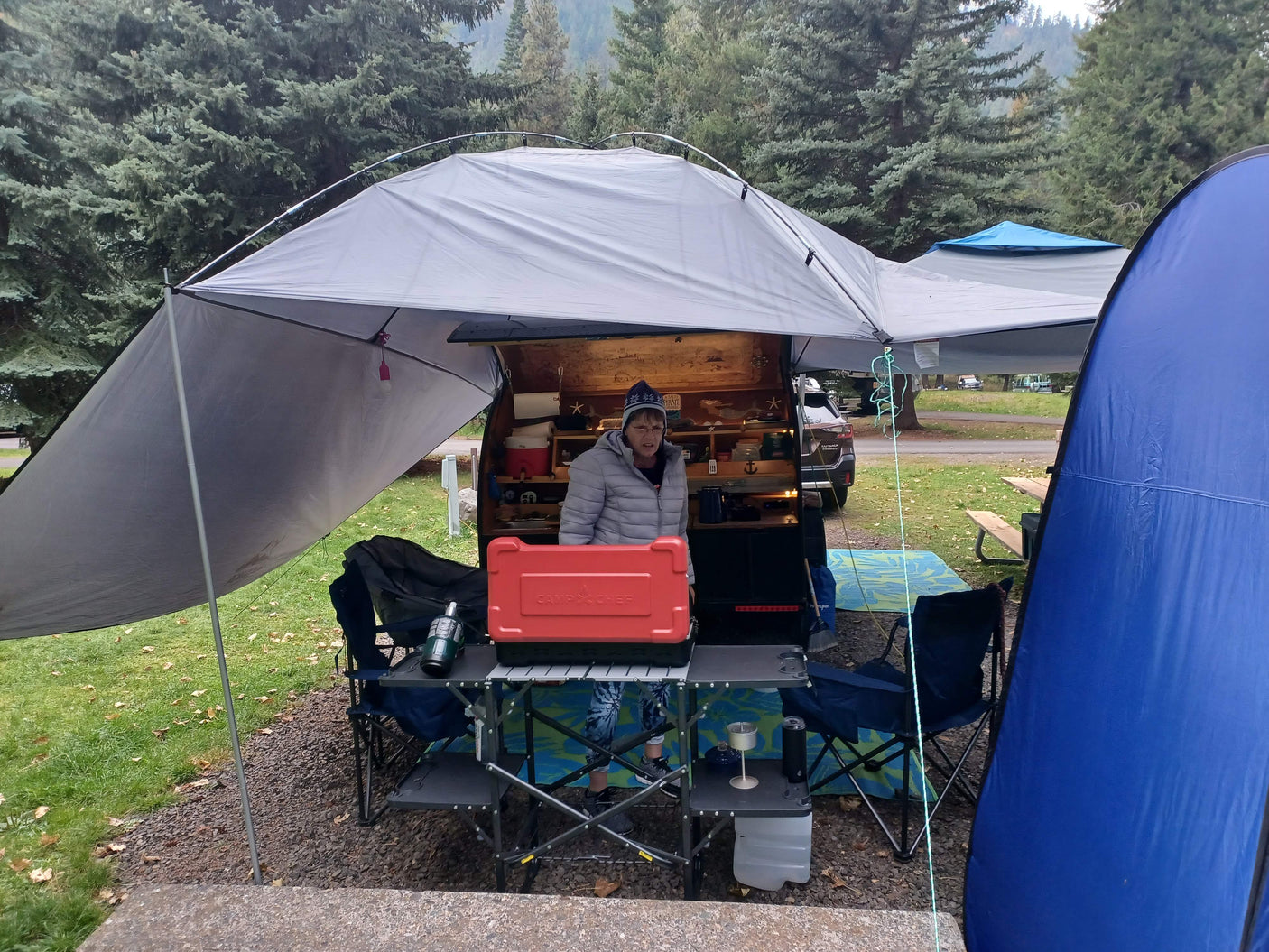 Woman cooking at an outdoor kitchen with a teardrop RV rental, sheltered by a large canopy at a scenic campground.