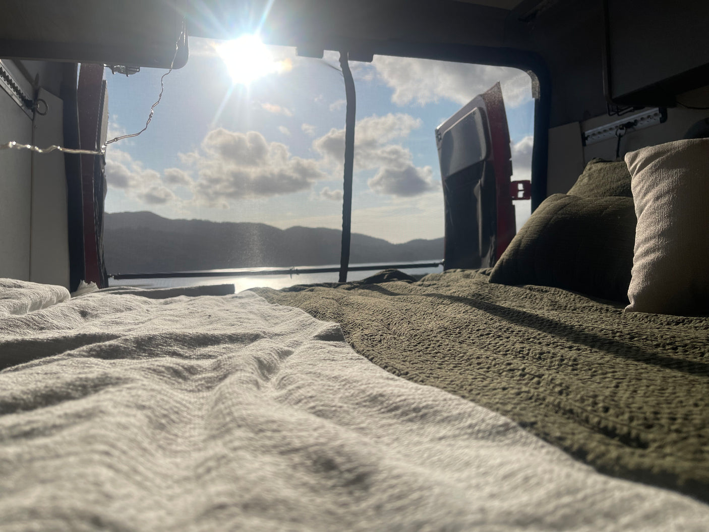An inviting bed in an rv rental, showcasing a beautiful serene lake and mountain view through the open rear doors on a sunny day.