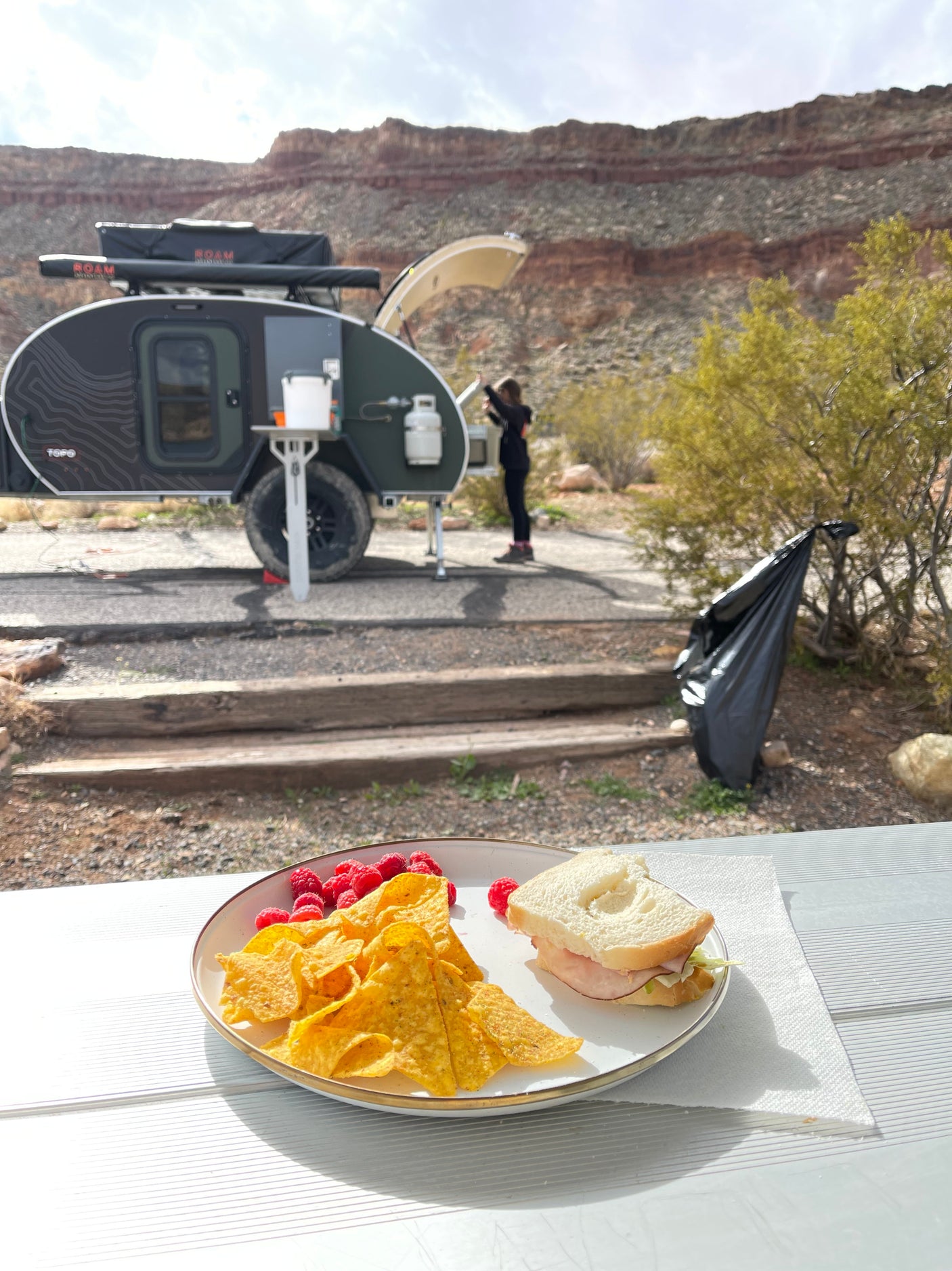 Outdoor meal at a beautiful RV rental campsite. Features a teardrop camper, a person, and scenic desert cliffs.