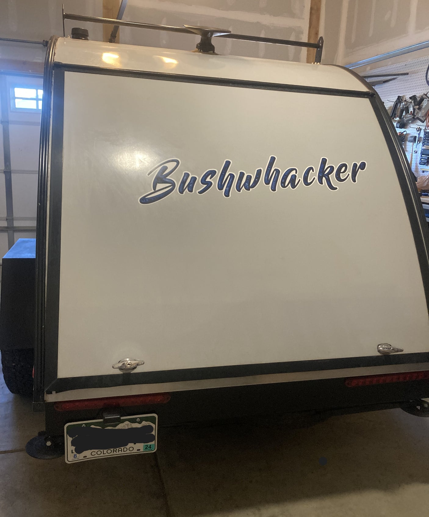Rear of a white Bushwhacker travel trailer (RV rental option) in a garage, with a roof rack & Colorado plate.