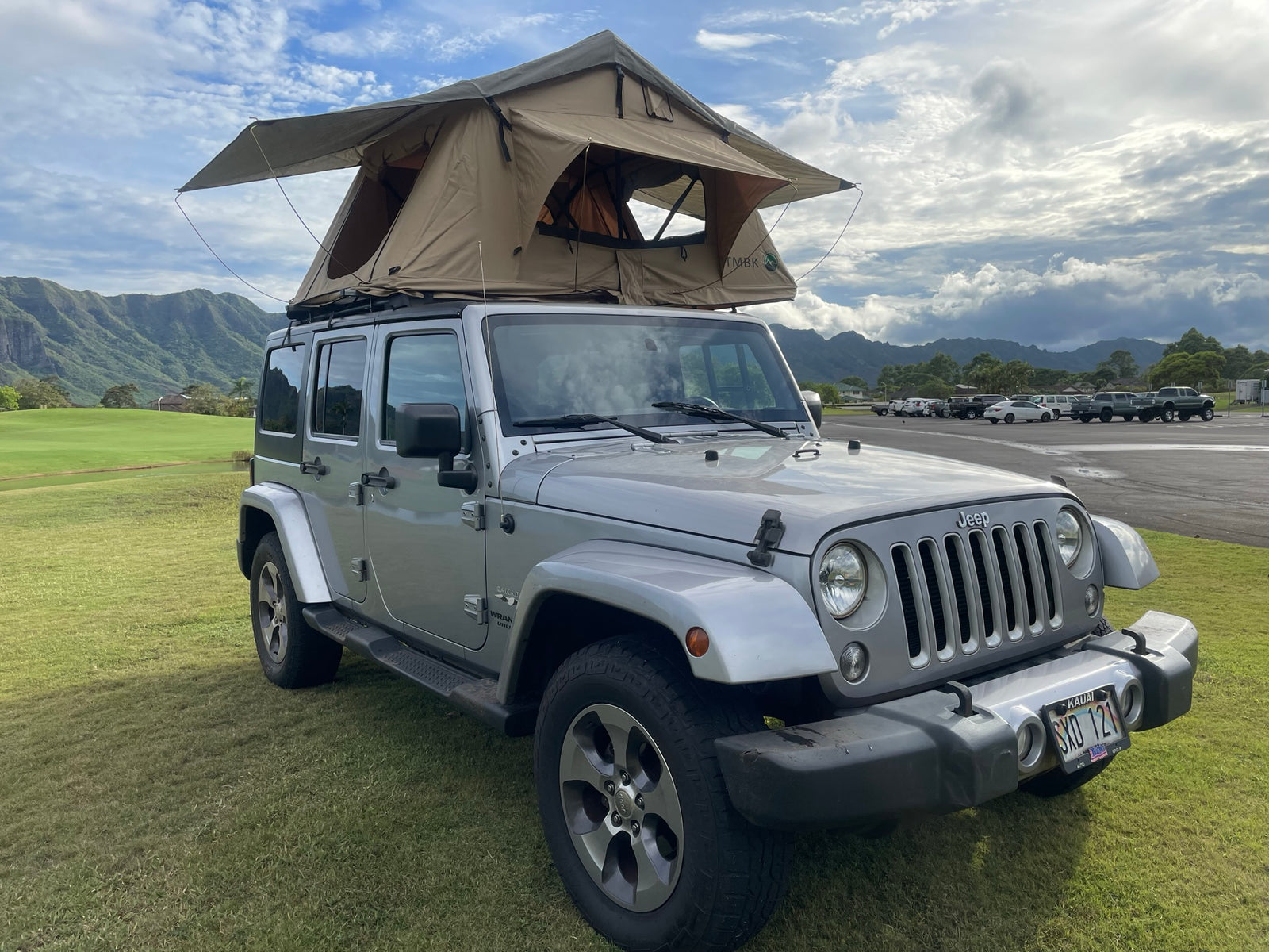 Silver Jeep Wrangler with a rooftop tent on green grass in Kauai. An ideal rv rental for outdoor adventures near mountains.