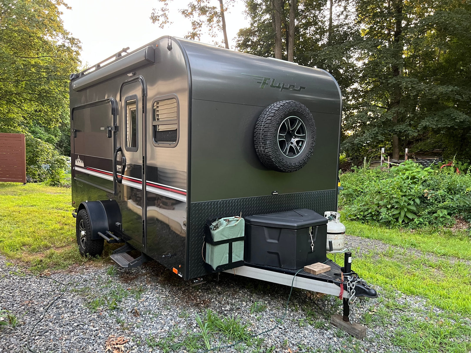 Dark green "Flyer" camper, perfect for an RV rental adventure, parked on gravel amidst trees and grass.