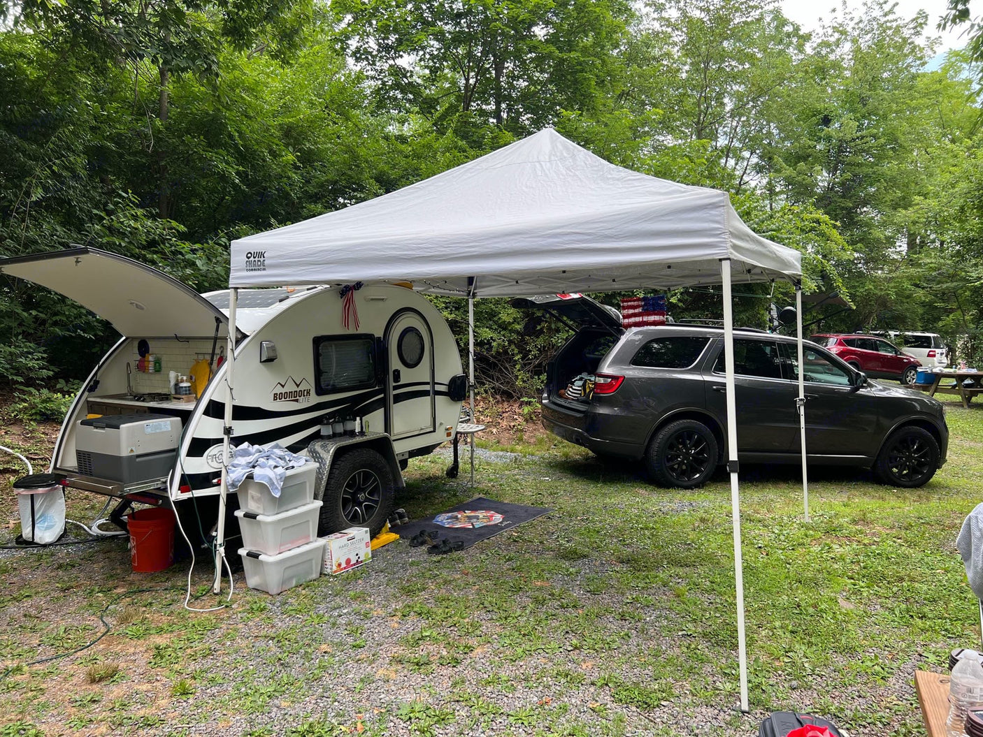 with chairs & pet bed.|White SUV towing a teardrop RV rental at a wooded campsite with gravel