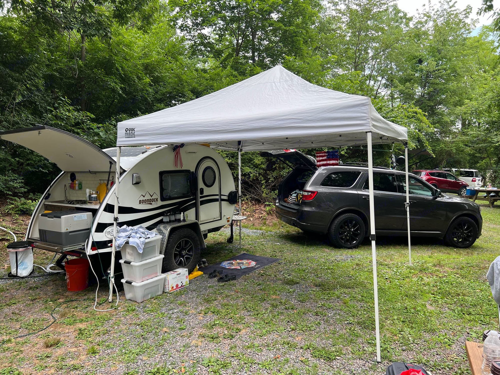 with chairs & pet bed.|White SUV towing a teardrop RV rental at a wooded campsite with gravel