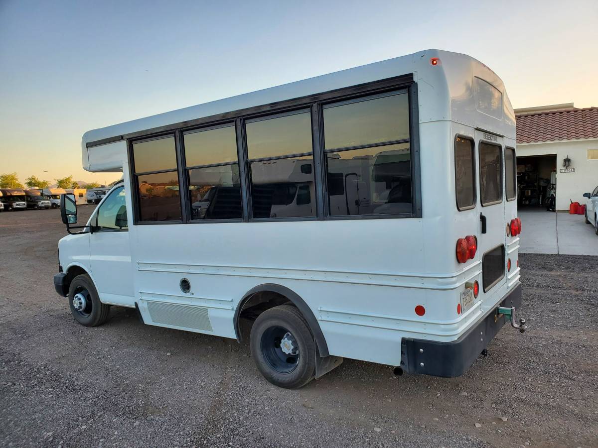 White shuttle bus, potential RV rental conversion, parked on gravel. Tinted windows, red tail lights, and tow hitch visible. — photo 5