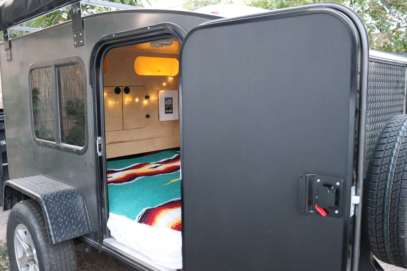 Open door view of a cozy teardrop camper RV rental interior, featuring warm wooden walls, string lights, and colorful bedding.