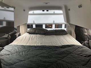 Inside an RV rental, a wide bed with gray bedding offers a comfy sleeping area for your camper van adventures.