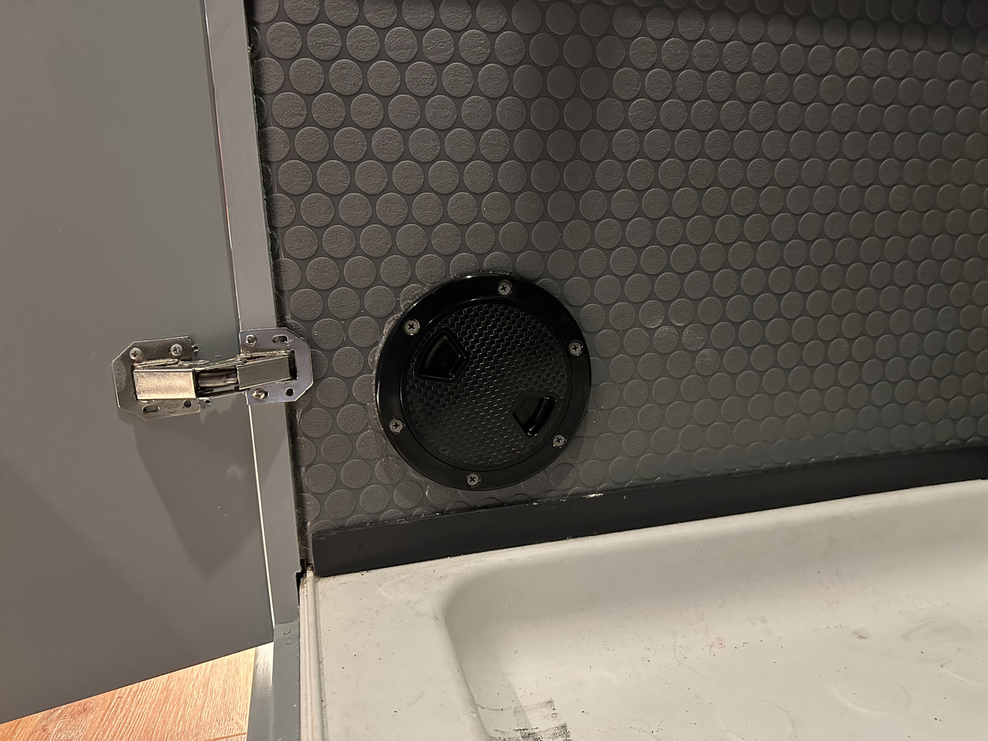 RV rental interior: Grey door with hinge, dark grey textured wall, black access hatch, and white sink below.